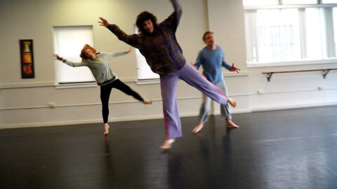 Three people rehearsing in a dance studio jumping up on an angle with their arms in the air.