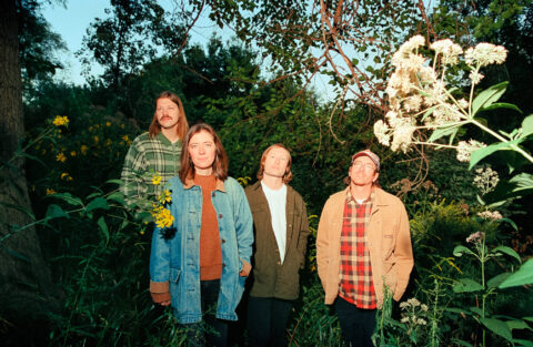 The four members of the band Ratboys wear autumnal dress and stand in a verdant field