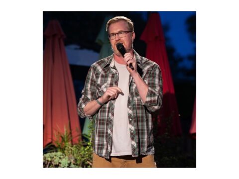 Comedian Kurt Braunohler wears a plaid shirt and holds a microphone