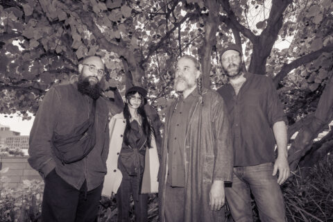 The four members of jazz group Natural Information Society stand near a tree with flowers in bloom in this black and white photo