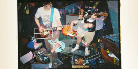 In a multi-exposure film shot, electric guitarists gaze at their fretboards and swirl about over the backdrop of a dimly lit stage crowded with pedals.