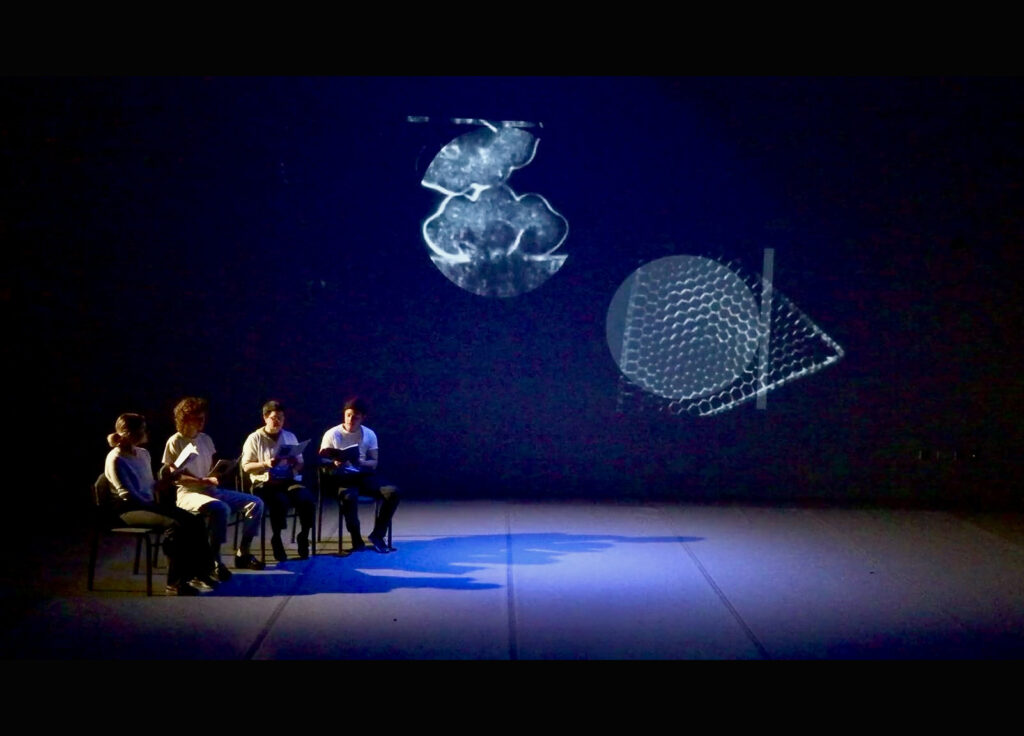 A photograph of a screening-performance of the work with microbial images projected and four figures reading from a songbook while seated.