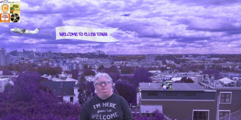 An individual with short grey hair and glasses, wearing a black shirt that reads, "I'm here, you're welcome," stands in front of a city under a purple haze. There is a drumset on a rooftop, and a plane flying by with a banner that reads, "Welcome to Ellen Town."