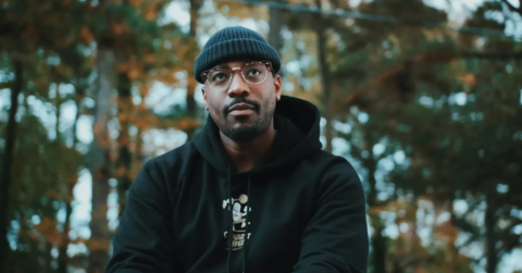 Tre Charles wearing a beanie and glasses in a black hoodie in the woods.