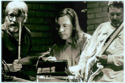 Close up on three guys; one is at a drumset, the other at an anolog synth, and the third playing bass very high up the neck.