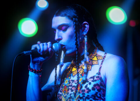 A photograph of Bobby Sanchez with a microphone performing on stage with blue lights.