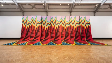 A brightly colored abstract geometric textile work by Liz Collins draping off the wall of the gallery and spilling onto the floor.