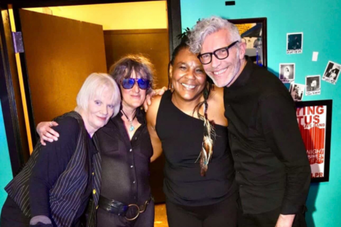The four members of the band Bush Tetras embrace while posing for a photograph in a small colorful room.